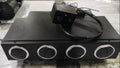 A black rectangular electronic control box with four circular silver-rimmed ports on the front, placed on a light gray marble-like surface. A small black wired control module sits on top of the unit.