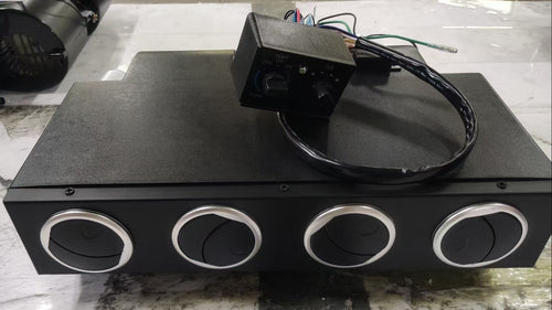 A black rectangular electronic control box with four circular silver-rimmed ports on the front, placed on a light gray marble-like surface. A small black wired control module sits on top of the unit.