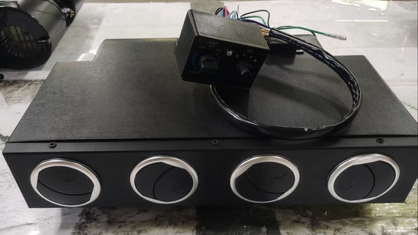 A black rectangular electronic control box with four circular silver-rimmed ports on the front, placed on a light gray marble-like surface. A small black wired control module sits on top of the unit.